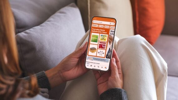 A person sitting on a couch uses a smartphone to browse an online grocery delivery app, displaying various grocery items and delivery options on the screen.
