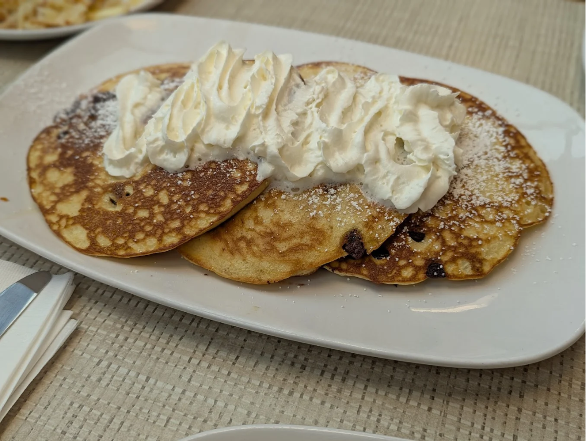 Three pancakes topped with powdered sugar and a generous swirl of whipped cream are served on a white rectangular plate, placed on a woven beige placemat.