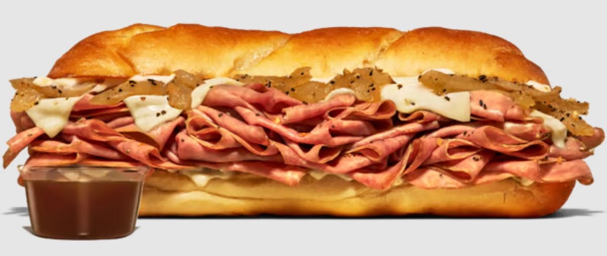A new fast-food menu classic: a sub sandwich with layers of thinly sliced roast beef, melted cheese, and grilled onions on a toasted roll, served with a small cup of brown dipping sauce.