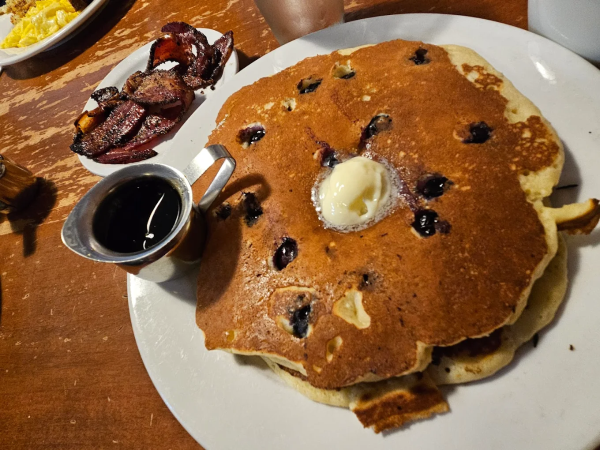 A large blueberry pancake with a pat of butter and a side of syrup on a white plate. Crispy bacon and scrambled eggs are visible in the background on a wooden table.