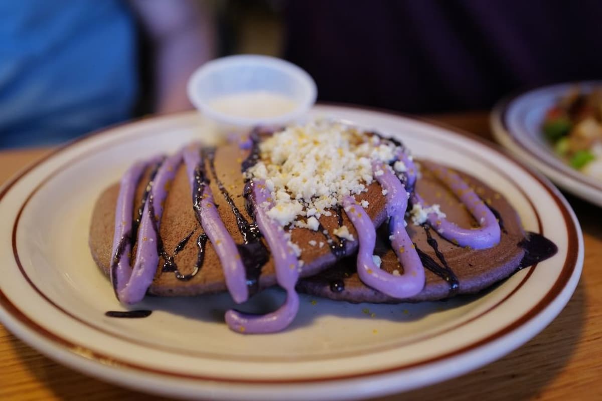 Three purple pancakes on a plate are topped with crumbled cheese, a drizzle of purple sauce, and a small cup of white sauce—worthy contenders for the best pancakes in every state.