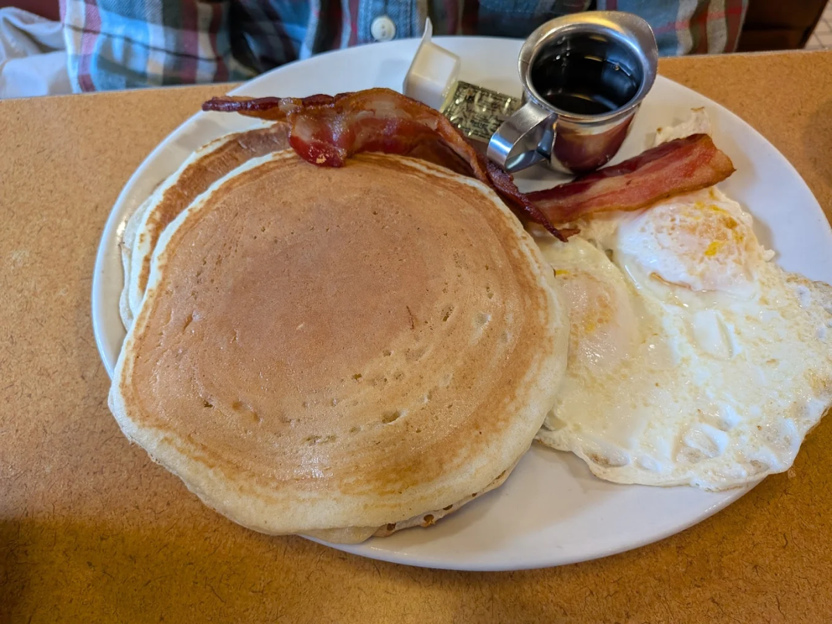 A breakfast plate with two pancakes, two strips of bacon, two fried eggs, a square of butter, a packet of syrup, and a small metal cup of syrup, set on a brown table. A person in a plaid shirt is in the background.
