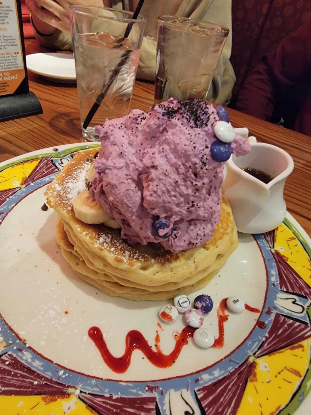 A stack of pancakes topped with banana slices, purple whipped cream, and colorful candy pieces sits on a decorated plate. A small pitcher of syrup and two glasses of water are on the table in the background.