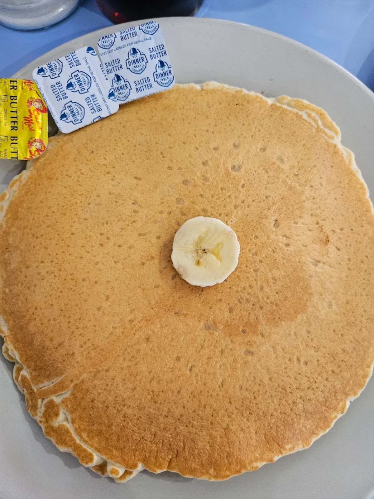 A large pancake on a white plate with a banana slice on top, served with packets of salted butter on the side.