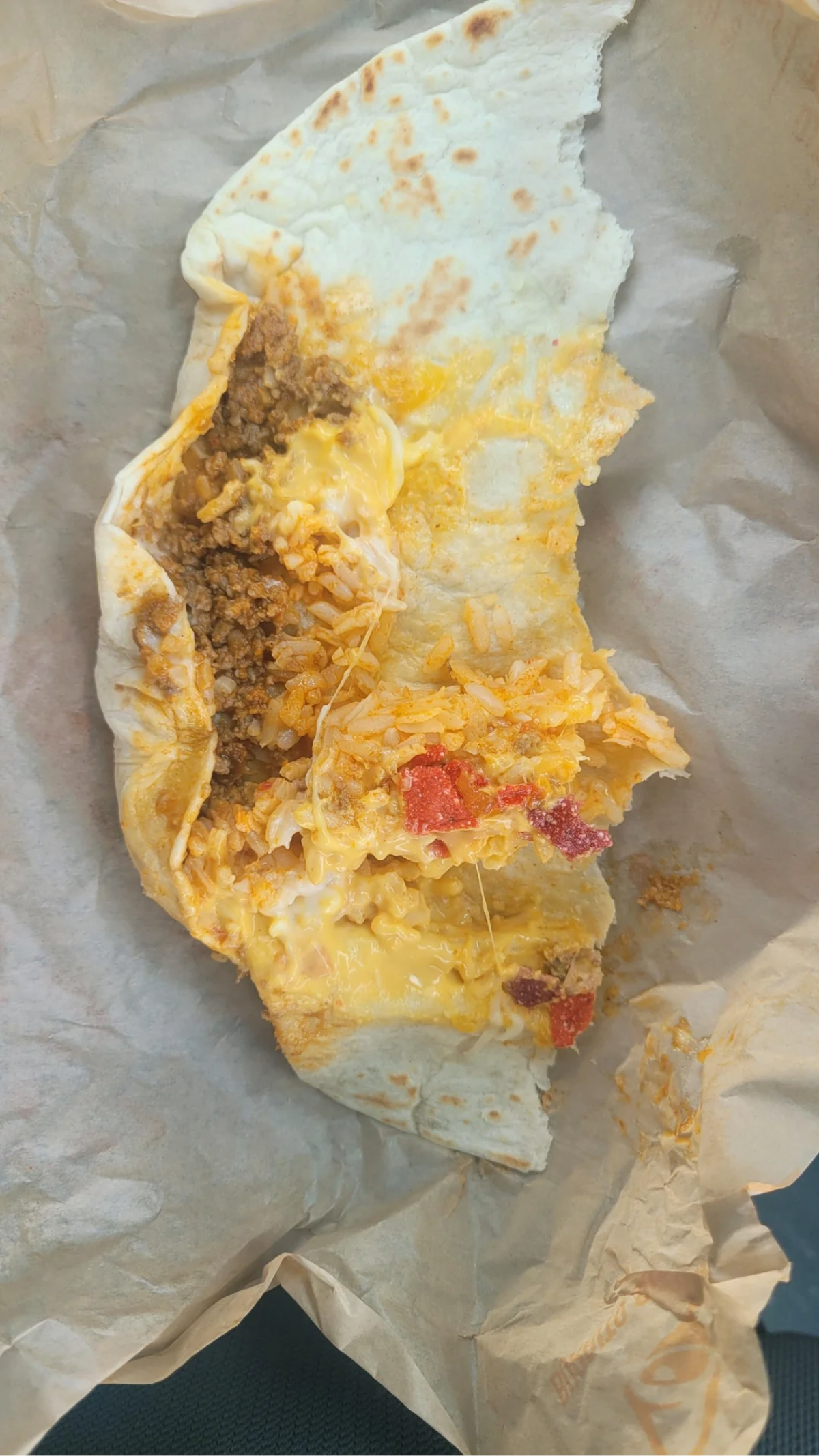 A partially unwrapped burrito on parchment paper, showing ground beef, rice, melted cheese, and pieces of tomato inside a flour tortilla.