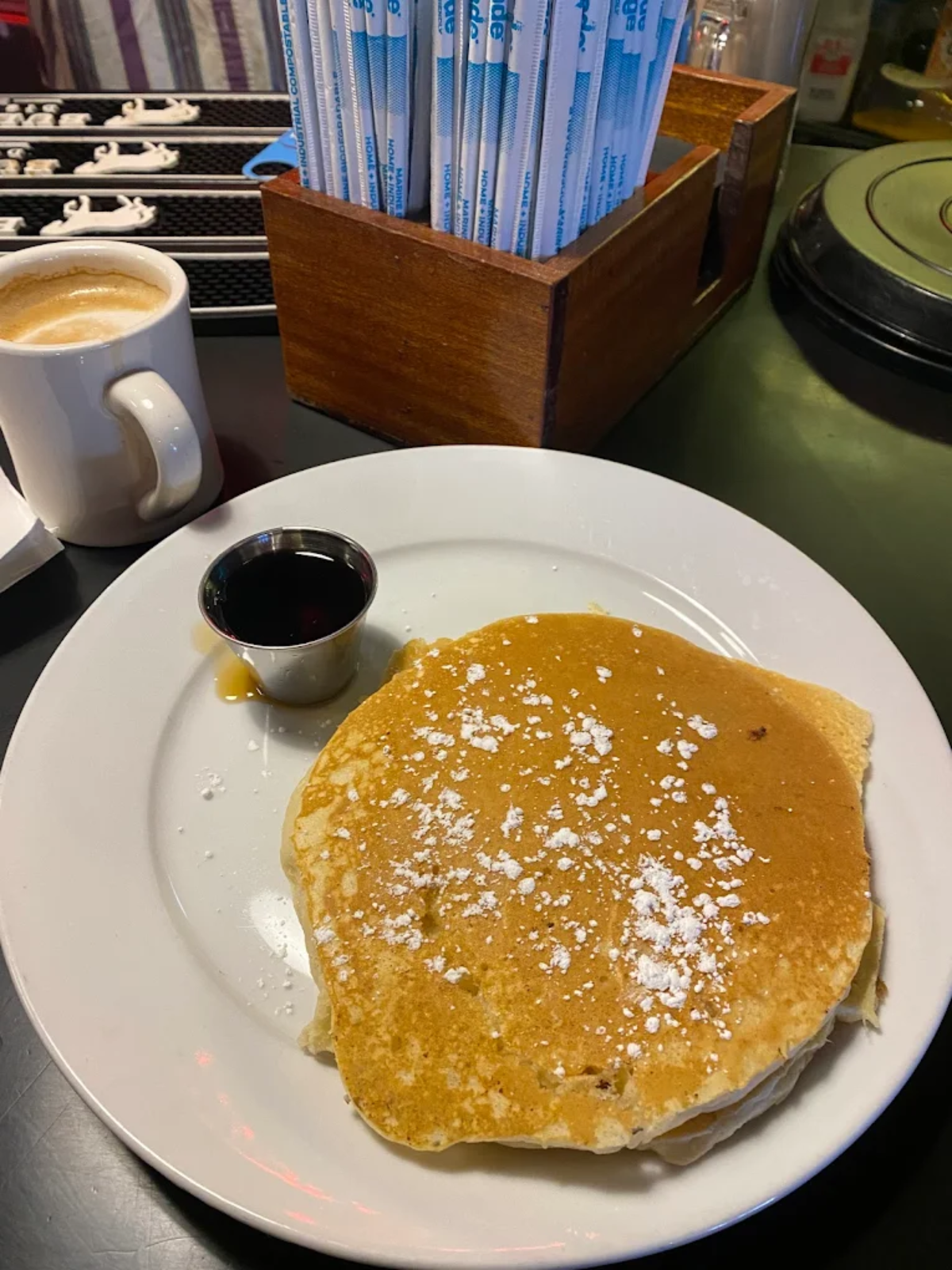 A plate with two pancakes topped with powdered sugar, a small metal cup of syrup, and a cup of coffee on the side. Behind them is a wooden box holding packets of sugar or straws.