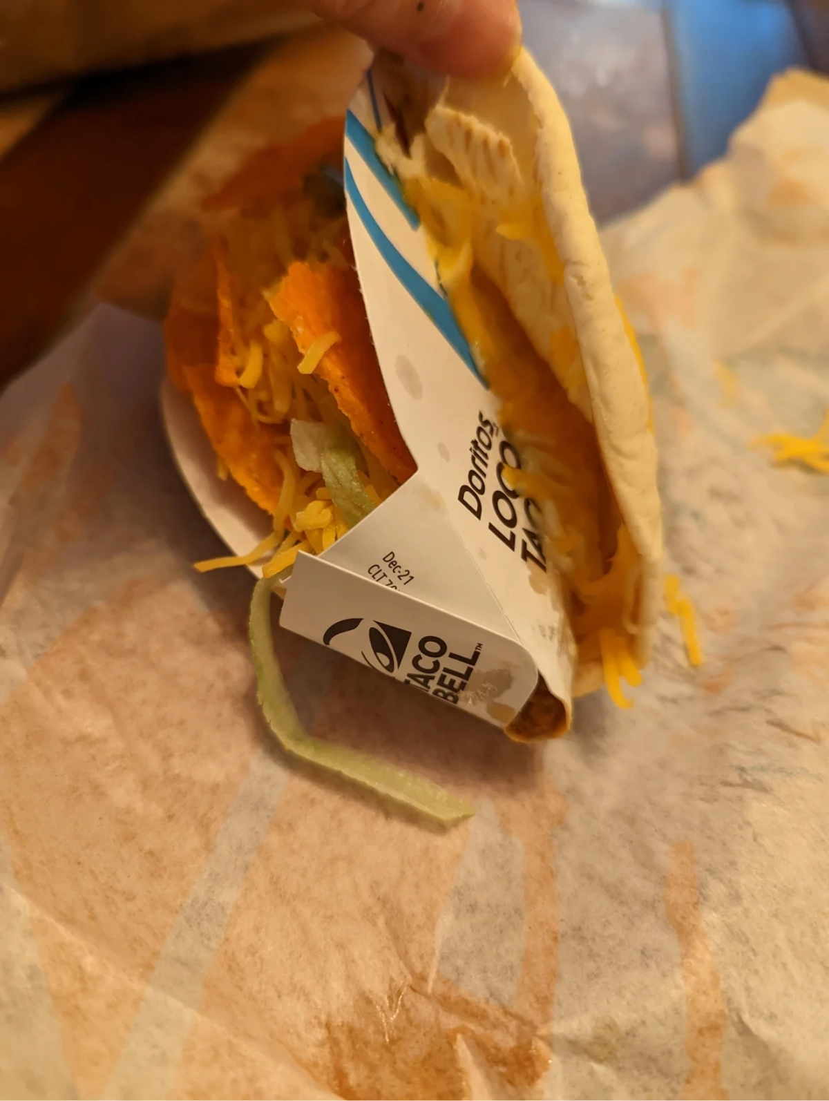 A partially unwrapped Taco Bell Doritos Locos Taco sits on a wrapper, revealing shredded cheese, lettuce, and the orange Doritos taco shell. A hand is slightly lifting the taco.