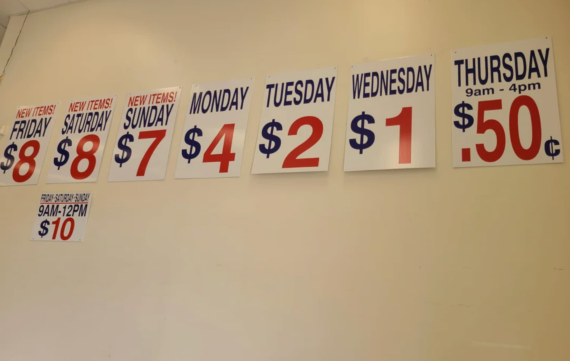 Seven signs on a wall show daily prices: Friday and Saturday $8, Sunday $7, Monday $4, Tuesday $2, Wednesday $1, Thursday 50¢. A smaller sign says "From Sunday-Sunday 9am-12pm $10.