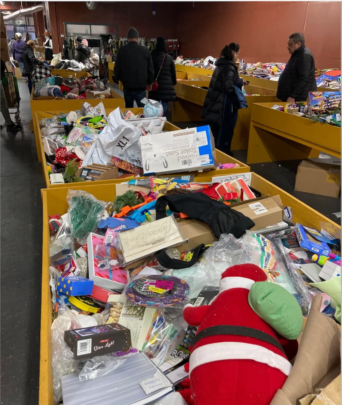 Shoppers browse large yellow bins filled with assorted toys, games, and miscellaneous items in a warehouse-style discount store. The background shows several people examining the goods.