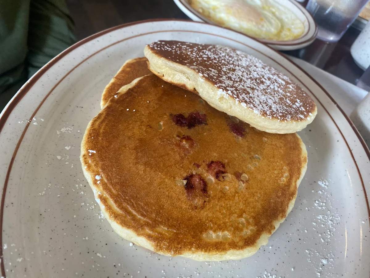 Two golden brown pancakes with visible berry pieces and a dusting of powdered sugar on a white plate with brown trim; another dish is blurred in the background.