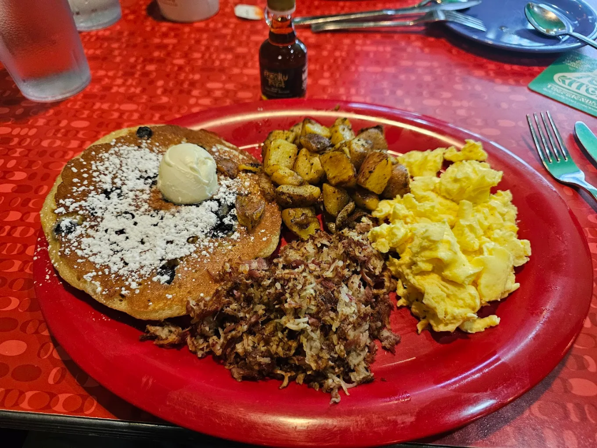 A red plate with scrambled eggs, seasoned breakfast potatoes, shredded corned beef hash, and a blueberry pancake topped with powdered sugar and a dollop of butter. A small bottle of syrup is in the background.