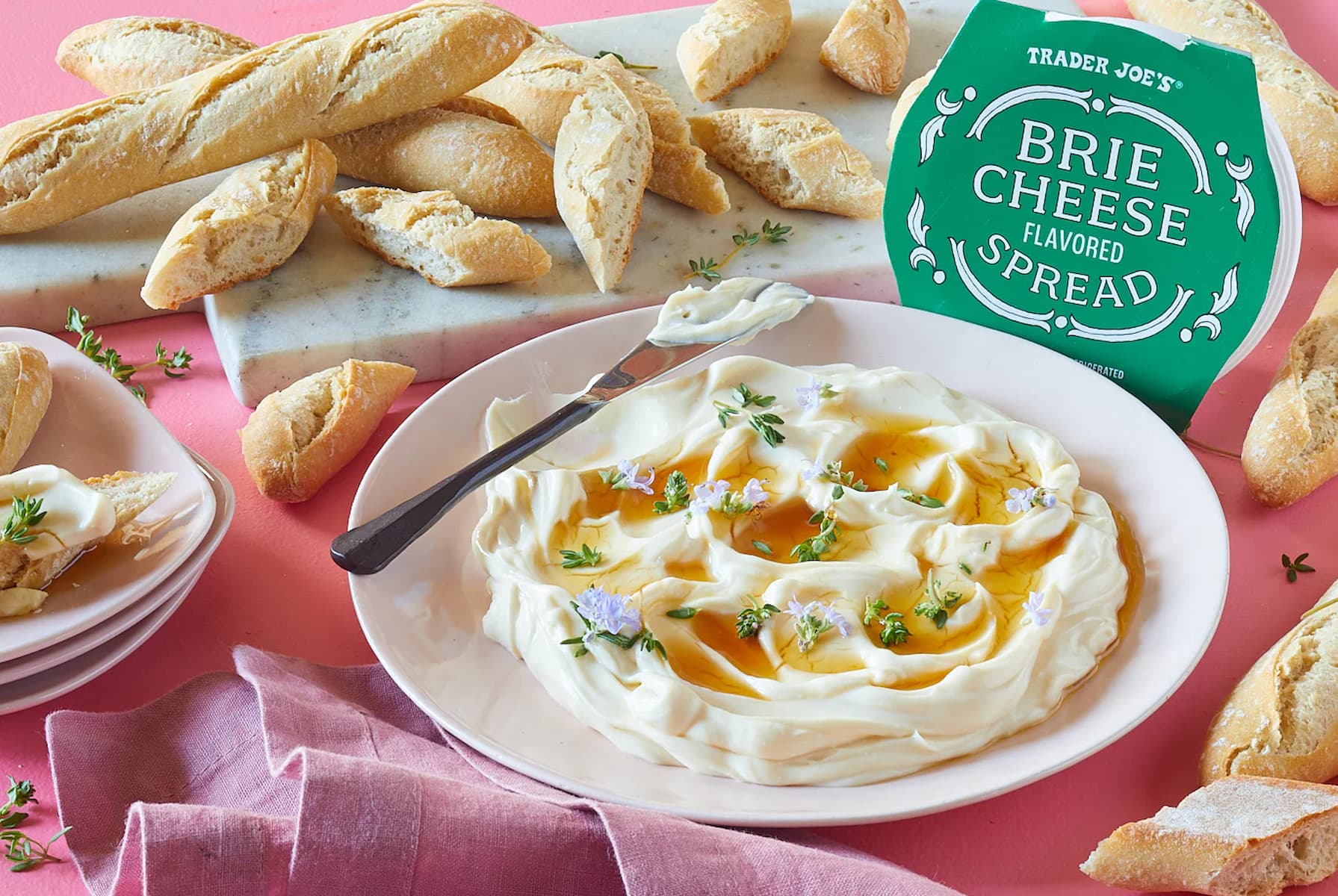 A plate of creamy brie cheese spread topped with herbs and honey, surrounded by sliced baguette pieces. A Trader Joe’s Brie Cheese Flavored Spread container is nearby on a pink surface with a purple napkin.