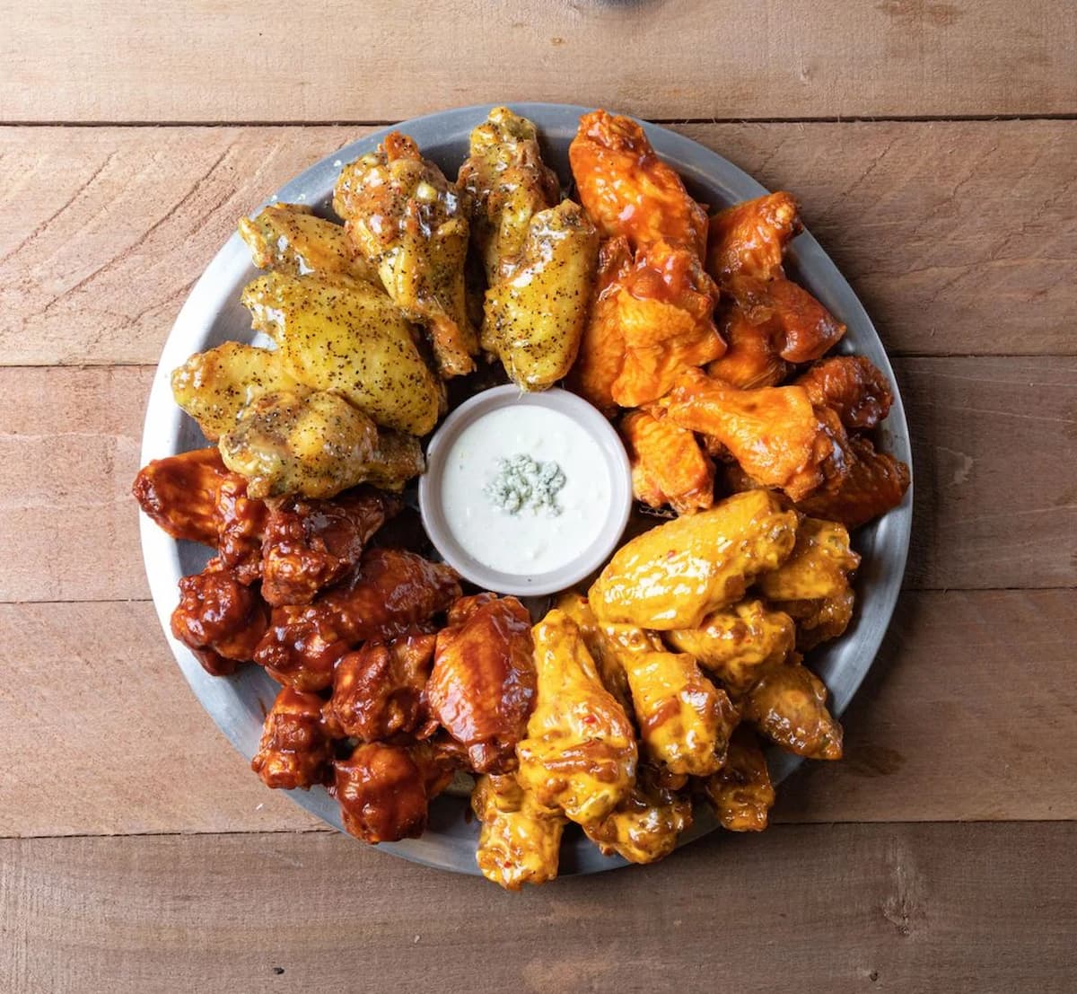 A round metal platter with four types of sauced chicken wings, perfect for sharing during March Madness restaurant deals, arranged in sections around a small cup of ranch dipping sauce and set on a wooden table.