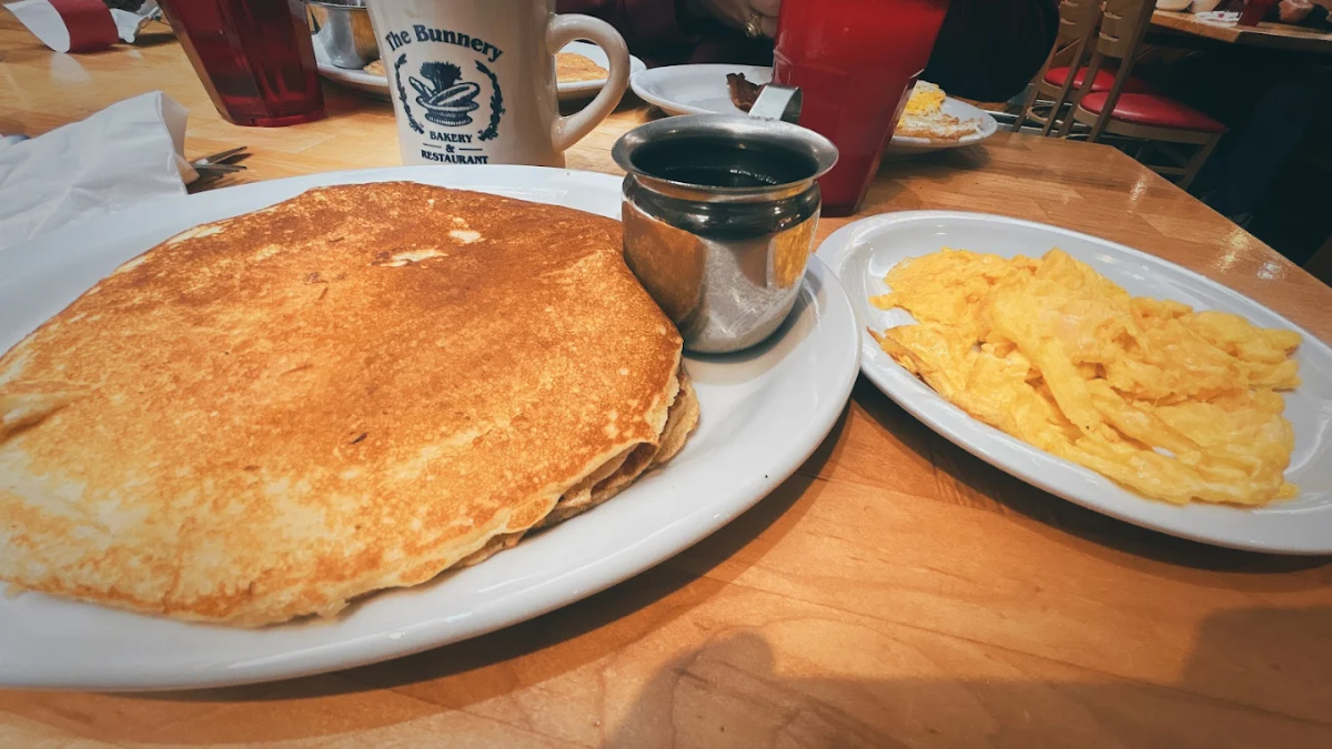 A breakfast plate with two large pancakes and a metal cup of syrup, next to a separate plate of scrambled eggs on a wooden table. A mug and red drink cups are in the background.