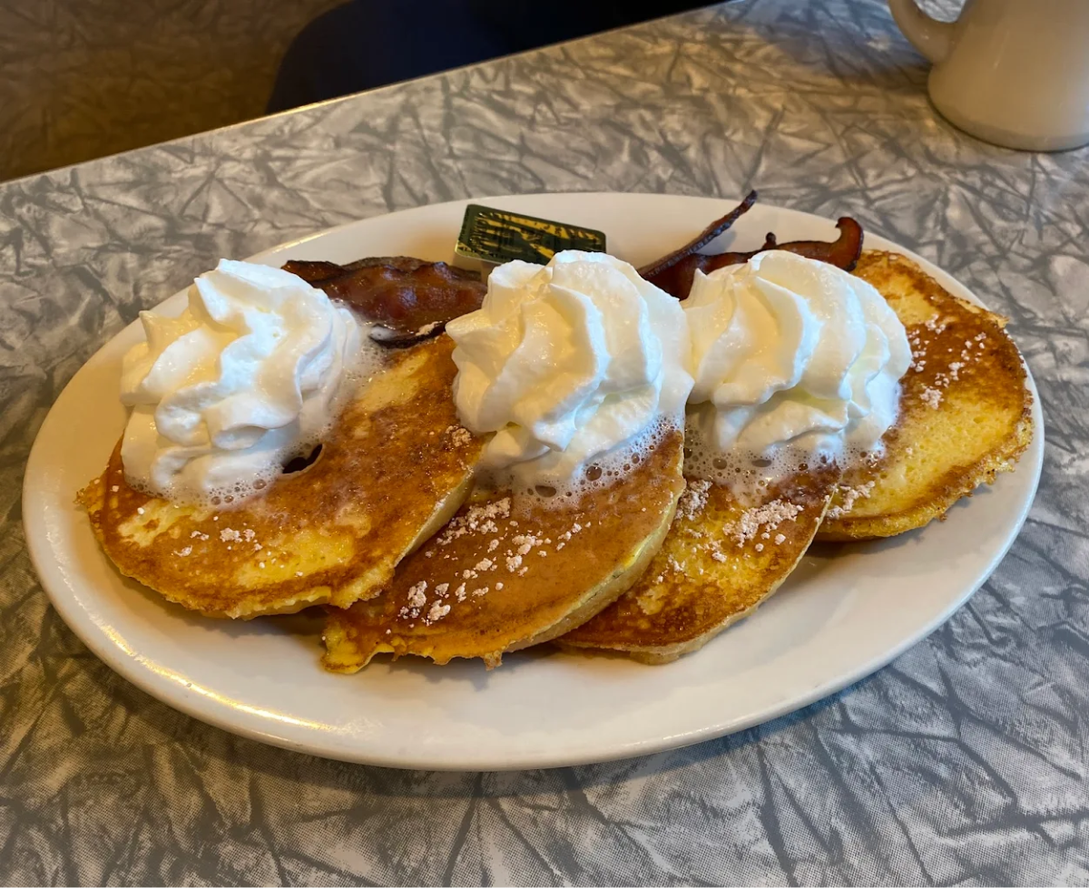 Three pancakes topped with whipped cream and powdered sugar are served on a white plate, accompanied by two strips of bacon, with a coffee mug in the background on a gray marbled table.