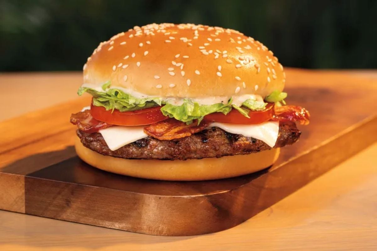 A cheeseburger with lettuce, tomato, bacon, and melted cheese in a sesame seed bun sits on a wooden platter, showcasing the new fast-food menu against a blurred background.