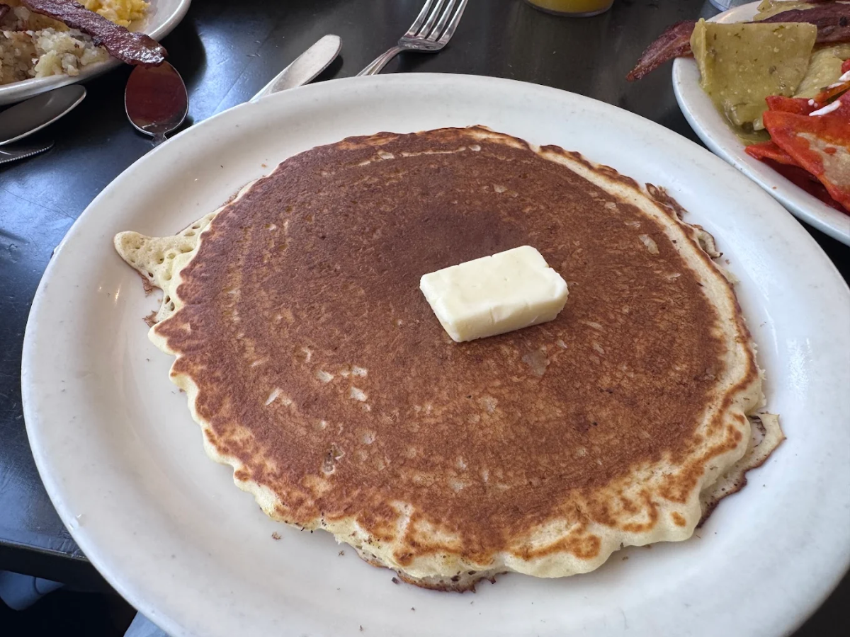 A large, golden-brown pancake with a pat of butter on top sits on a white plate, surrounded by breakfast foods including potatoes, bacon, and eggs. A fork and spoon are nearby on the table.