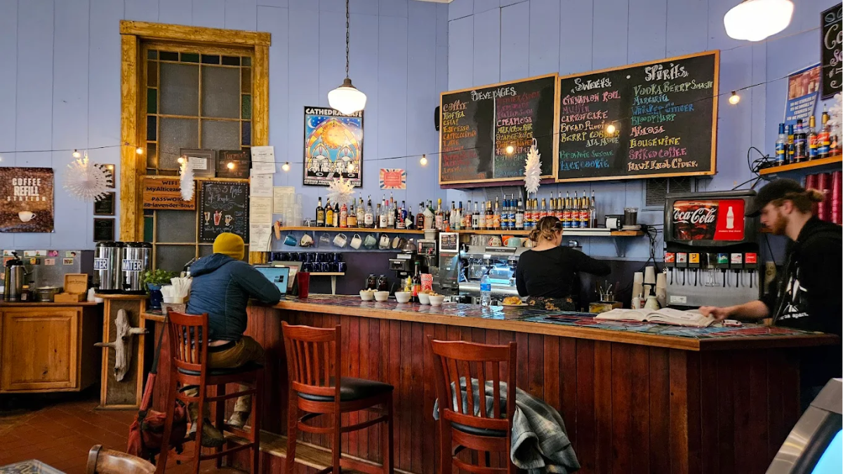 A cozy café with a wooden counter, colorful chalkboard menus, bottles on shelves, and warm lighting. A person in a yellow beanie uses a laptop at the bar while two staff members work behind the counter.