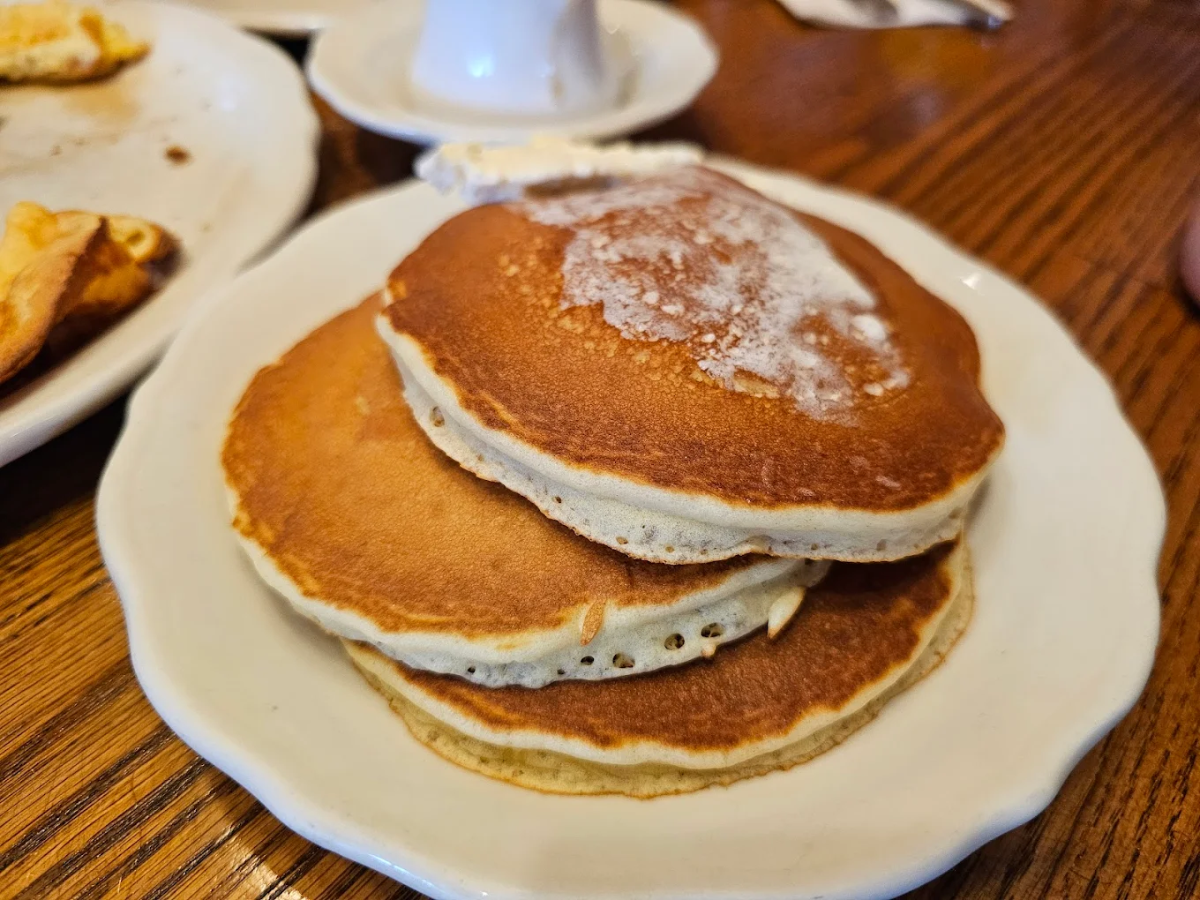 Three golden brown pancakes with a pat of melting butter on top, served on a white plate on a wooden table. Nearby, there is a partial view of another plate and a cup.