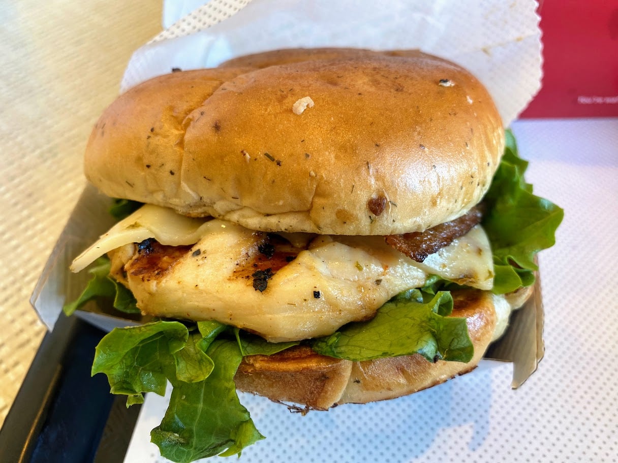 A grilled chicken sandwich with lettuce and melted cheese on a toasted bun, served in a paper container on a tray.