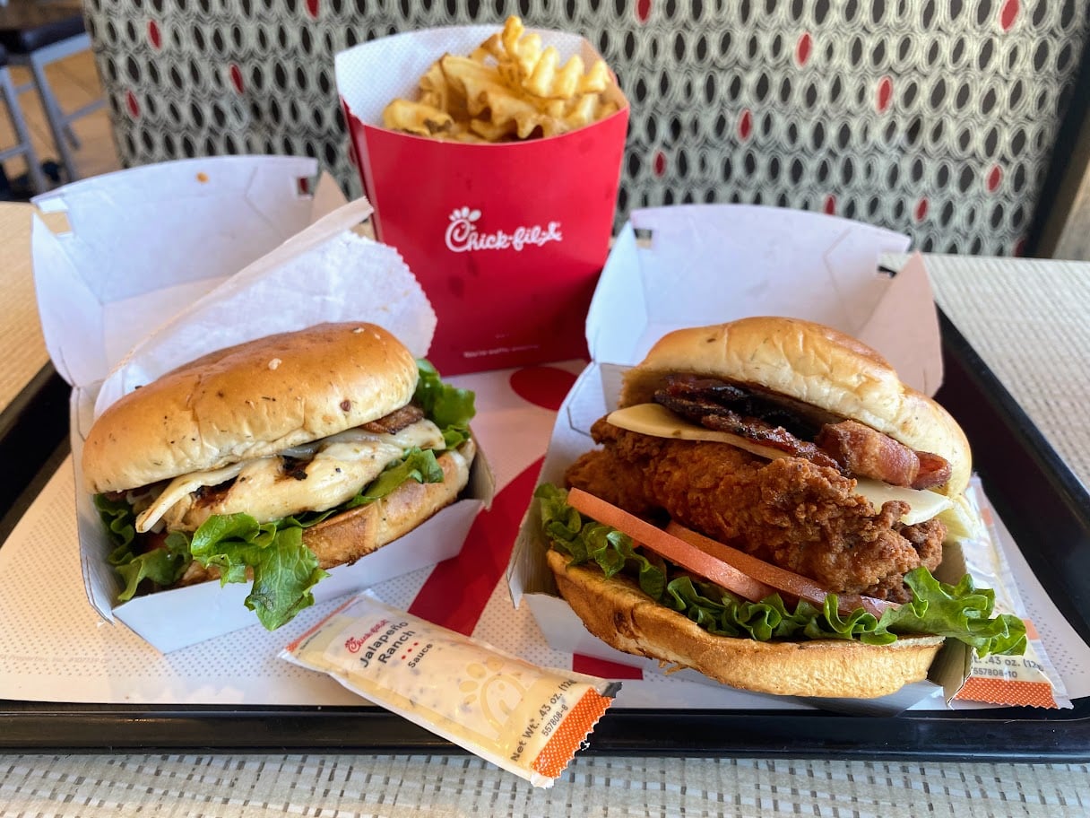 Two Chick-fil-A sandwiches—one grilled, one fried with bacon and cheese—are served with lettuce and tomato on a tray, alongside a box of waffle fries and a packet of Chick-fil-A sauce.