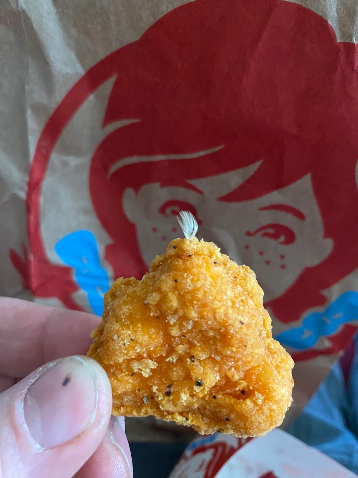 A hand holds a breaded chicken nugget with a small feather sticking out, in front of a Wendy’s paper bag featuring the restaurant’s red-haired logo.