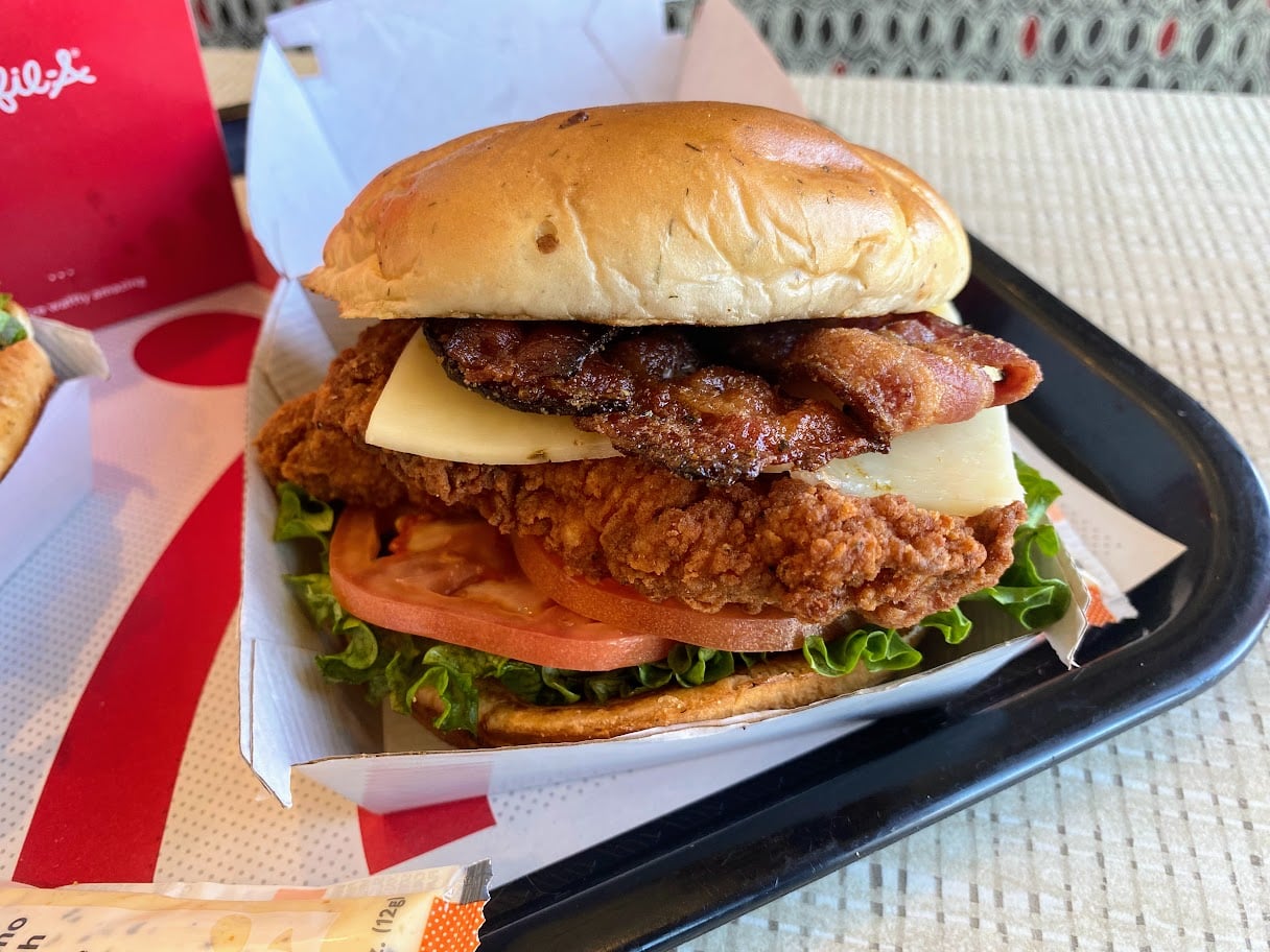 A crispy chicken sandwich with bacon, white cheese, lettuce, and tomato on a bun, served on a black tray with a red and white wrapper underneath.