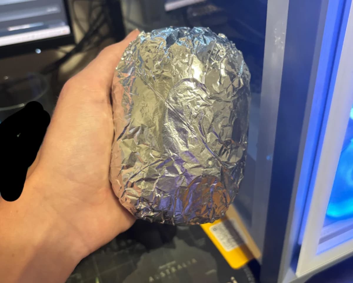 A hand is holding a large, oval-shaped object wrapped in crinkled aluminum foil—possibly the result of fast food fails—near a computer setup with visible monitors and a blue-lit PC case.
