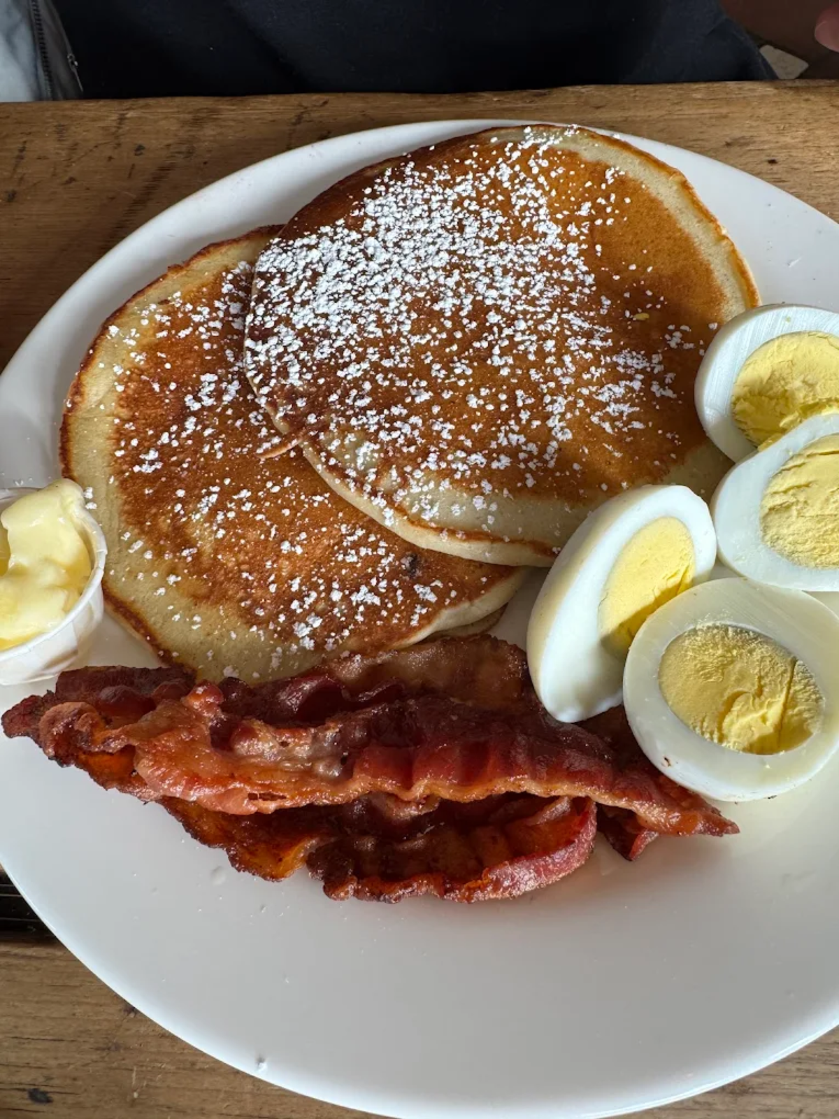 A plate with two pancakes topped with powdered sugar, sliced hard-boiled eggs, crispy bacon strips, and a small cup of butter on a wooden table.