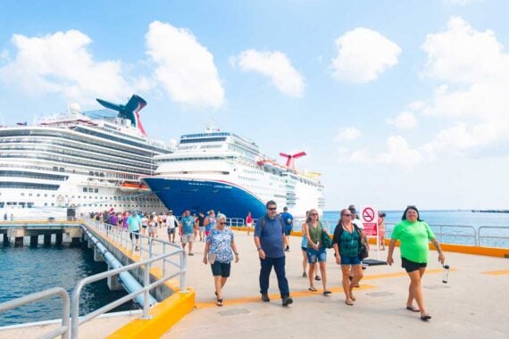 A group of people walk on a pier next to two large cruise ships docked under a bright, partly cloudy sky. Some, perhaps reflecting on cruise purchases not worth it, carry bags and wear casual summer clothing.