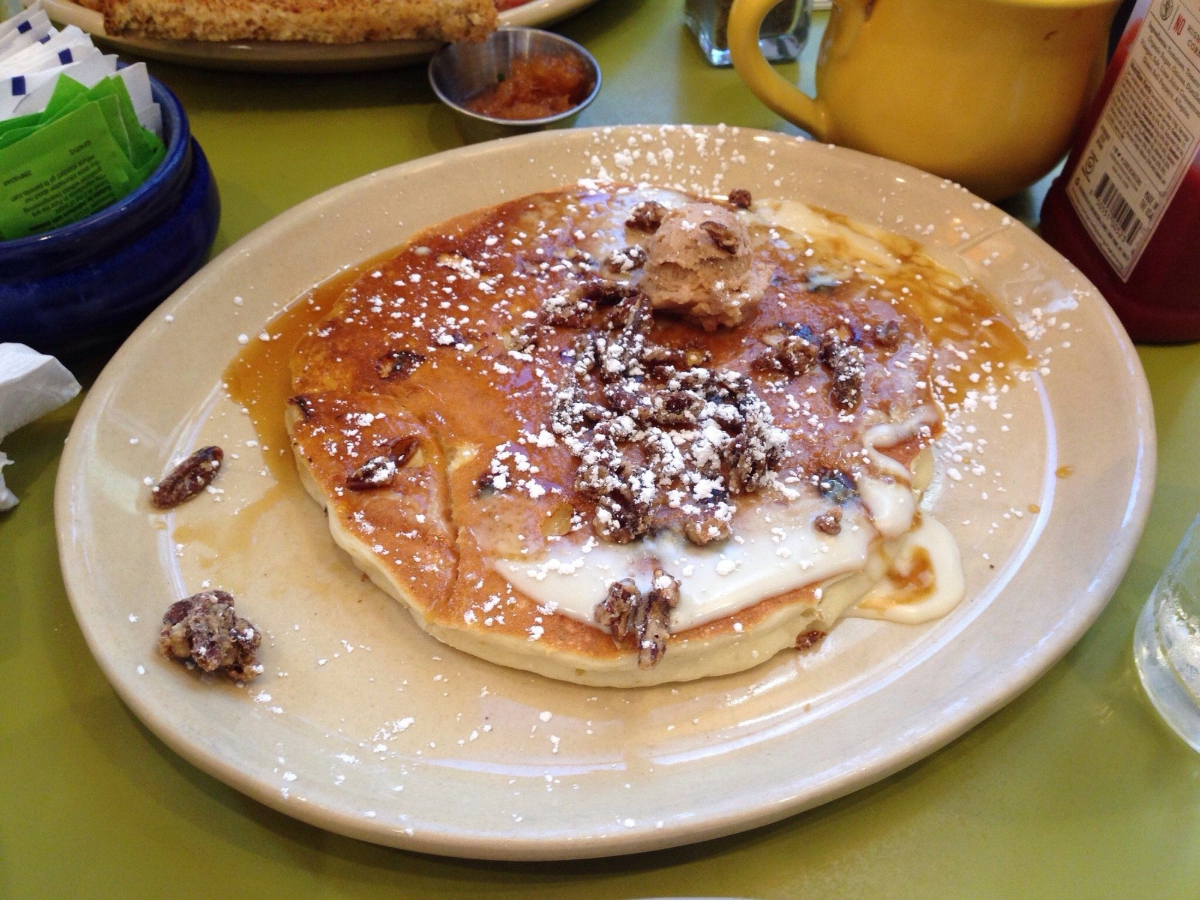 A plate of pancakes topped with powdered sugar, candied pecans, a dollop of cinnamon butter, and drizzled syrup sits on a green table with condiments and drinks in the background.