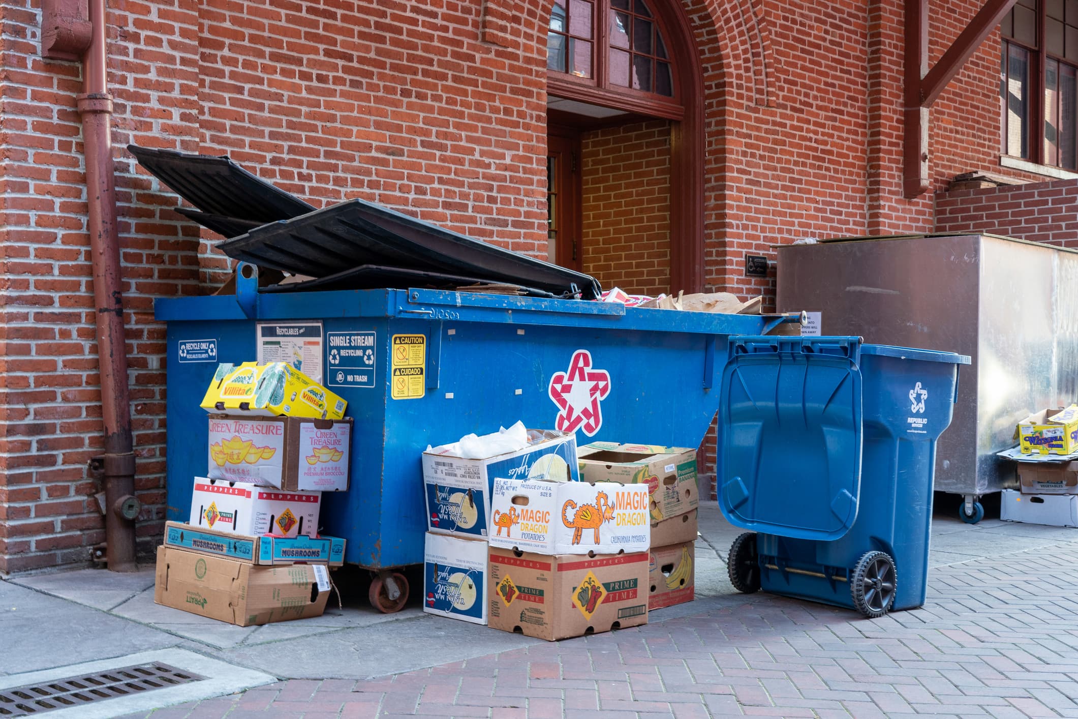 A large blue dumpster and recycling bin are surrounded by stacked cardboard boxes, some filled with discarded items, outside a brick building with arched windows and a metal pipe.