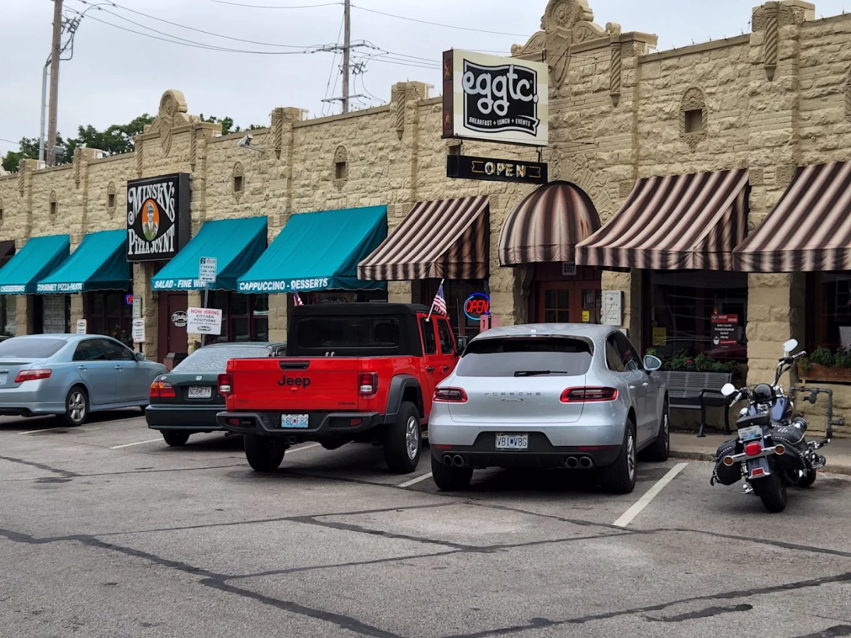 A restaurant with tan stone walls, striped awnings, and a sign reading "eggctc." Three cars and a motorcycle are parked in front, with a Guinness sign and blue awnings visible on the adjacent building.