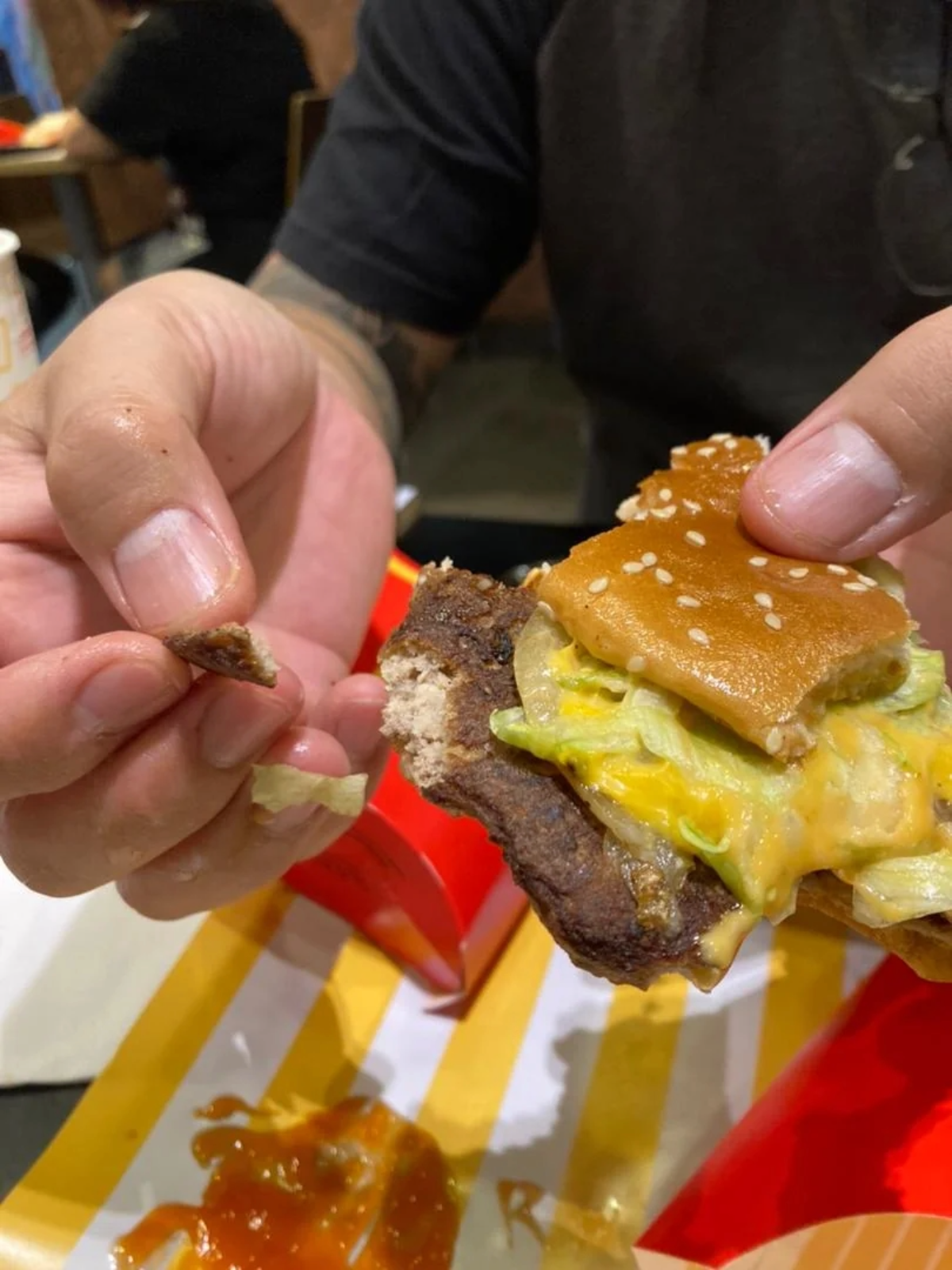 A person holding a partially eaten cheeseburger in one hand and a small piece of burger patty in the other, with a tray containing ketchup and an empty french fry container below.
