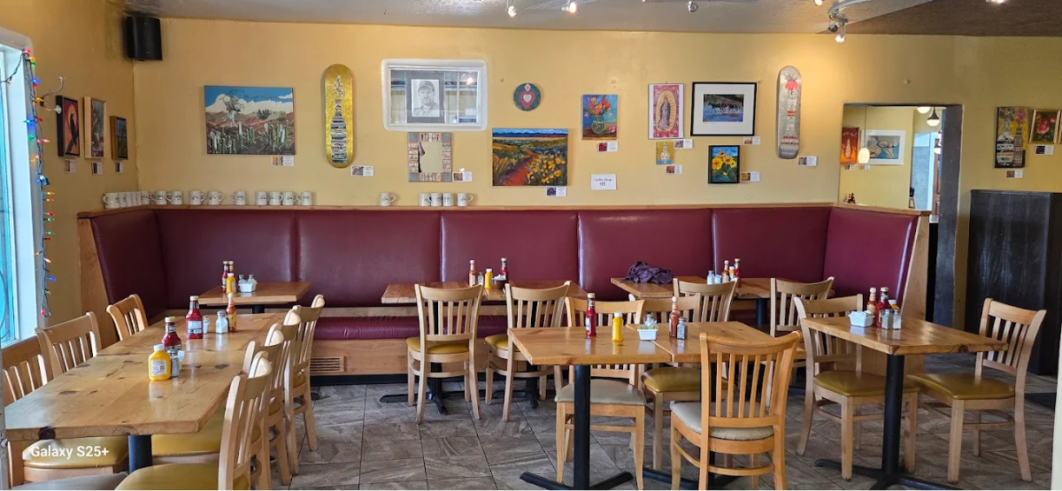 A cozy restaurant dining area with wooden tables and chairs, red bench seating, and many colorful paintings on yellow walls. Each table has condiments, and mugs line a shelf beneath the artwork.