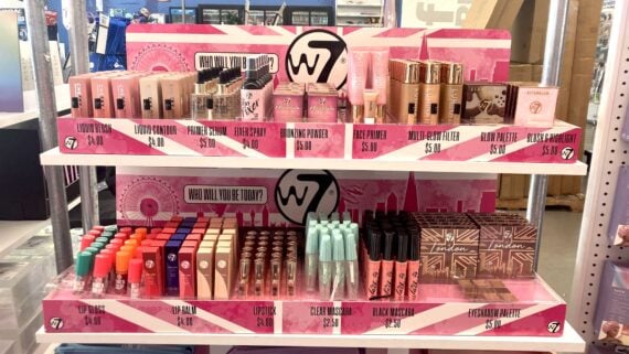 Two pink display shelves filled with W7 beauty products, including foundation, lipsticks, setting sprays, powders, highlighters, mascaras, and eyeshadow palettes&mdash;all neatly arranged with prices on the display.