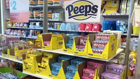 A store display features shelves stocked with various colorful Peeps marshmallow candy packages under a large Peeps sign, with pastel-colored Easter baskets and containers in the background.
