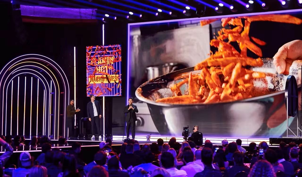 A person onstage speaks to an audience while a giant screen behind them displays a close-up of hands tossing sweet potato fries in a mixing bowl. Bright lights and colorful designs illuminate the stage.