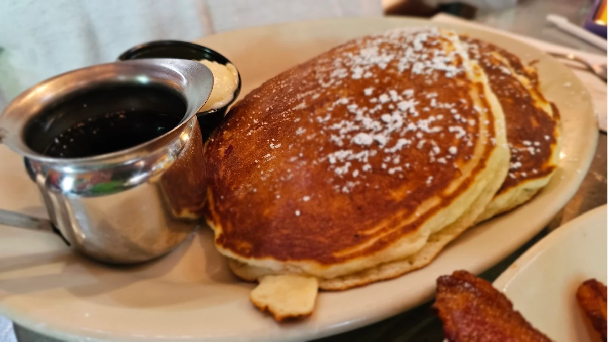 A plate with two large pancakes dusted with powdered sugar, served with a small metal pitcher of syrup and a cup of butter on the side. Part of a strip of bacon is visible in the foreground.