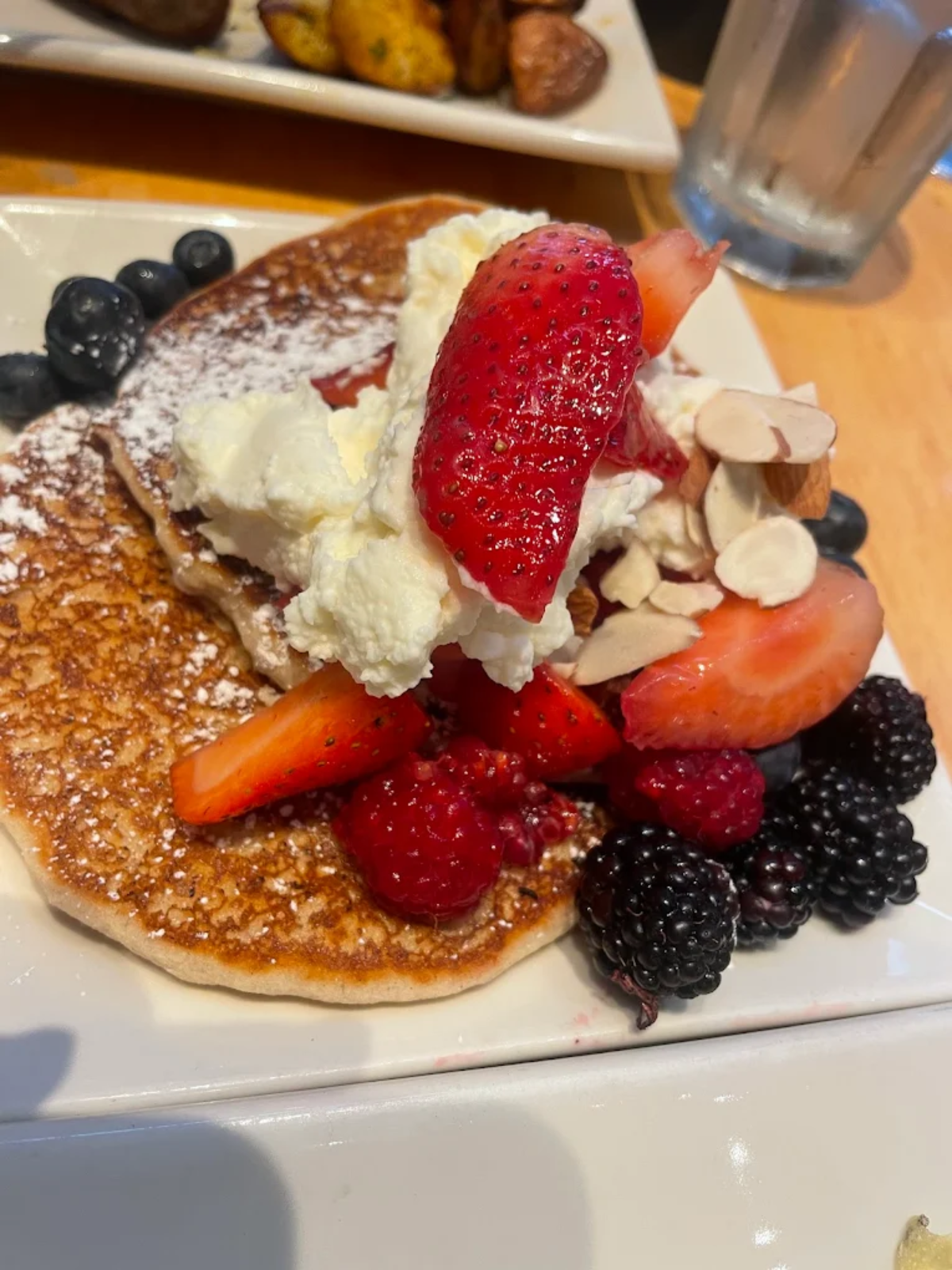 A plate with pancakes topped with whipped cream, sliced strawberries, blackberries, raspberries, blueberries, almond slices, and powdered sugar, with a glass of water and roasted potatoes in the background.
