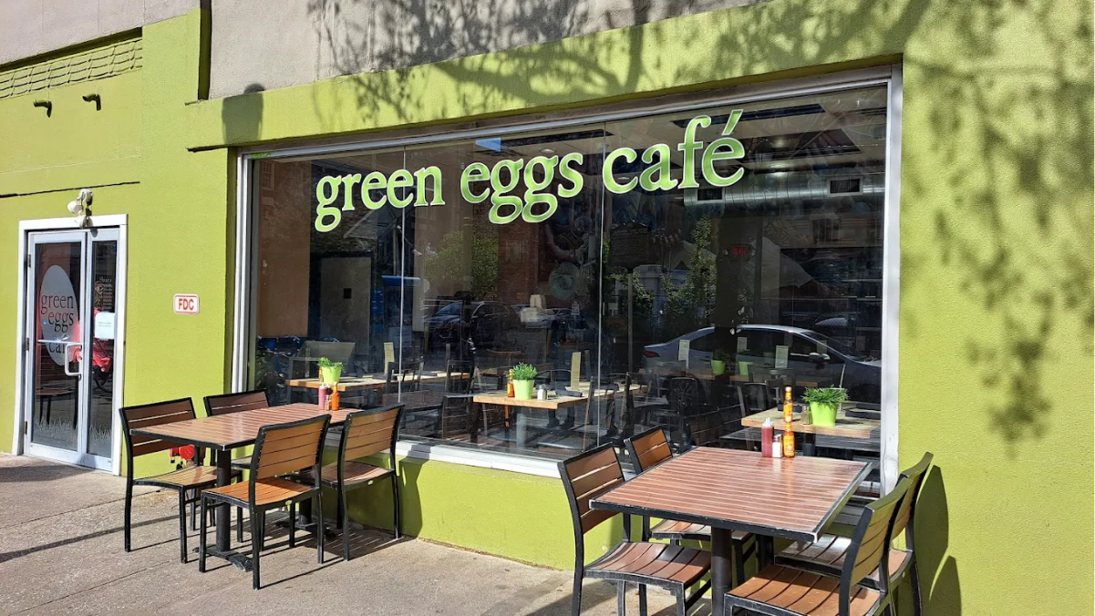 Green Eggs Café storefront with large windows, green exterior, and outdoor seating with wooden tables and chairs. Small potted plants sit on each table. The café name is displayed in large, light green letters on the window.