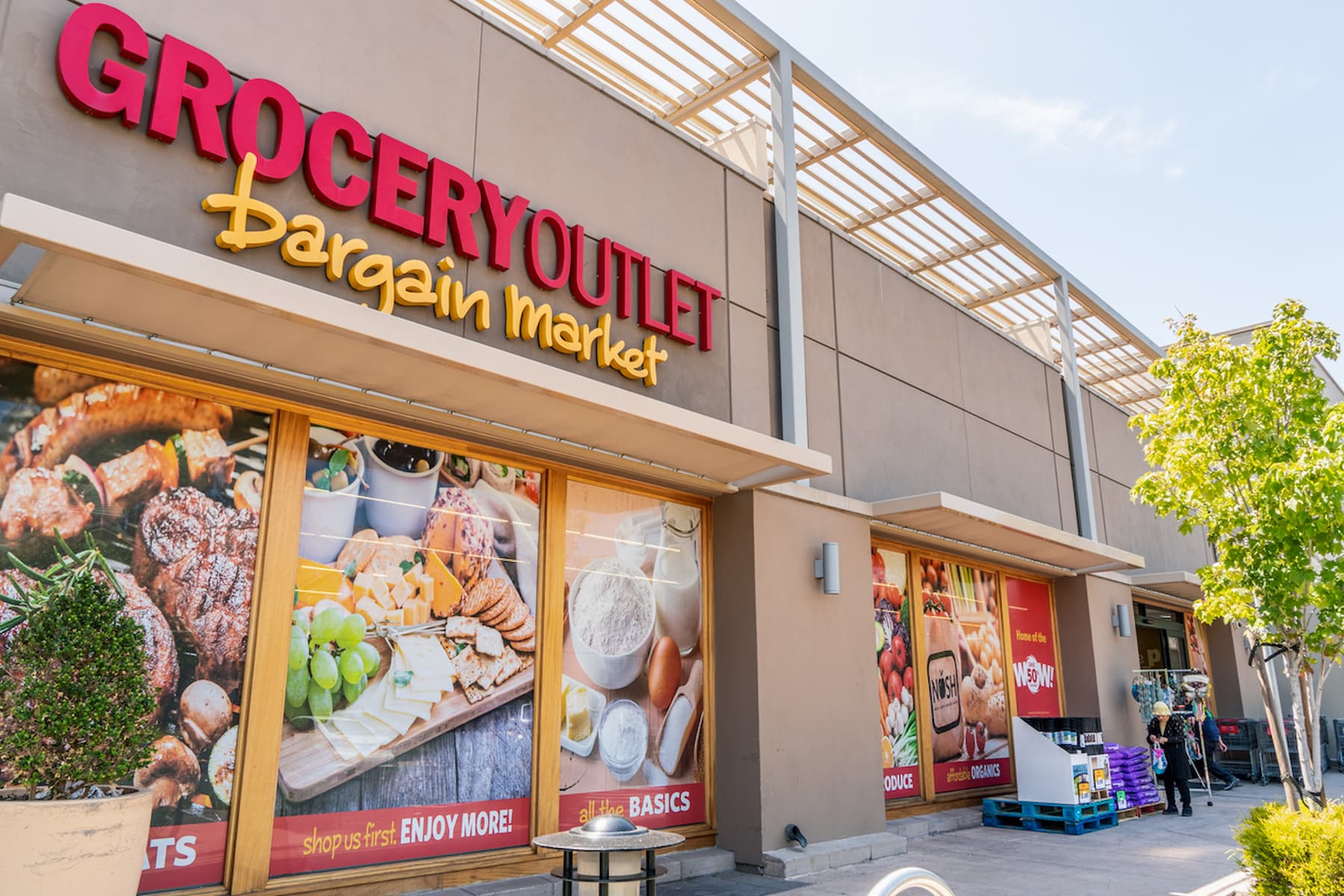 Exterior of a Grocery Outlet Bargain Market store, with large food images on the windows and a red and yellow sign above the entrance; a few people and shopping carts are near the entrance.