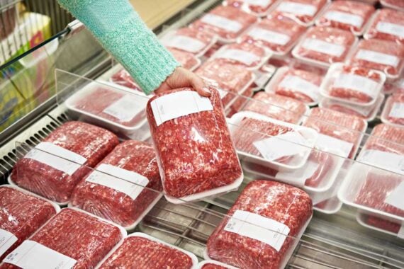 A person wearing a green sweater selects a package of ground meat from a refrigerated display filled with similar plastic-wrapped meat packages in a grocery store.
