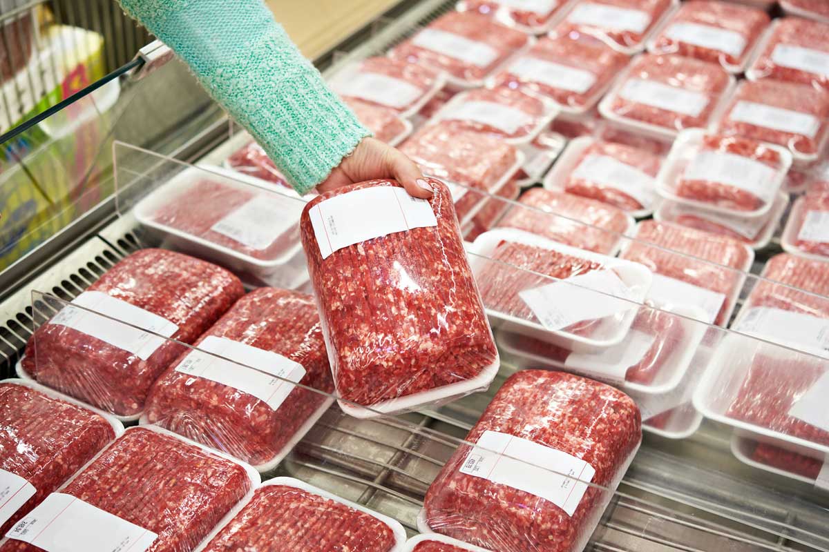 A person wearing a green sweater selects a package of ground meat from a refrigerated display filled with similar plastic-wrapped meat packages in a grocery store.