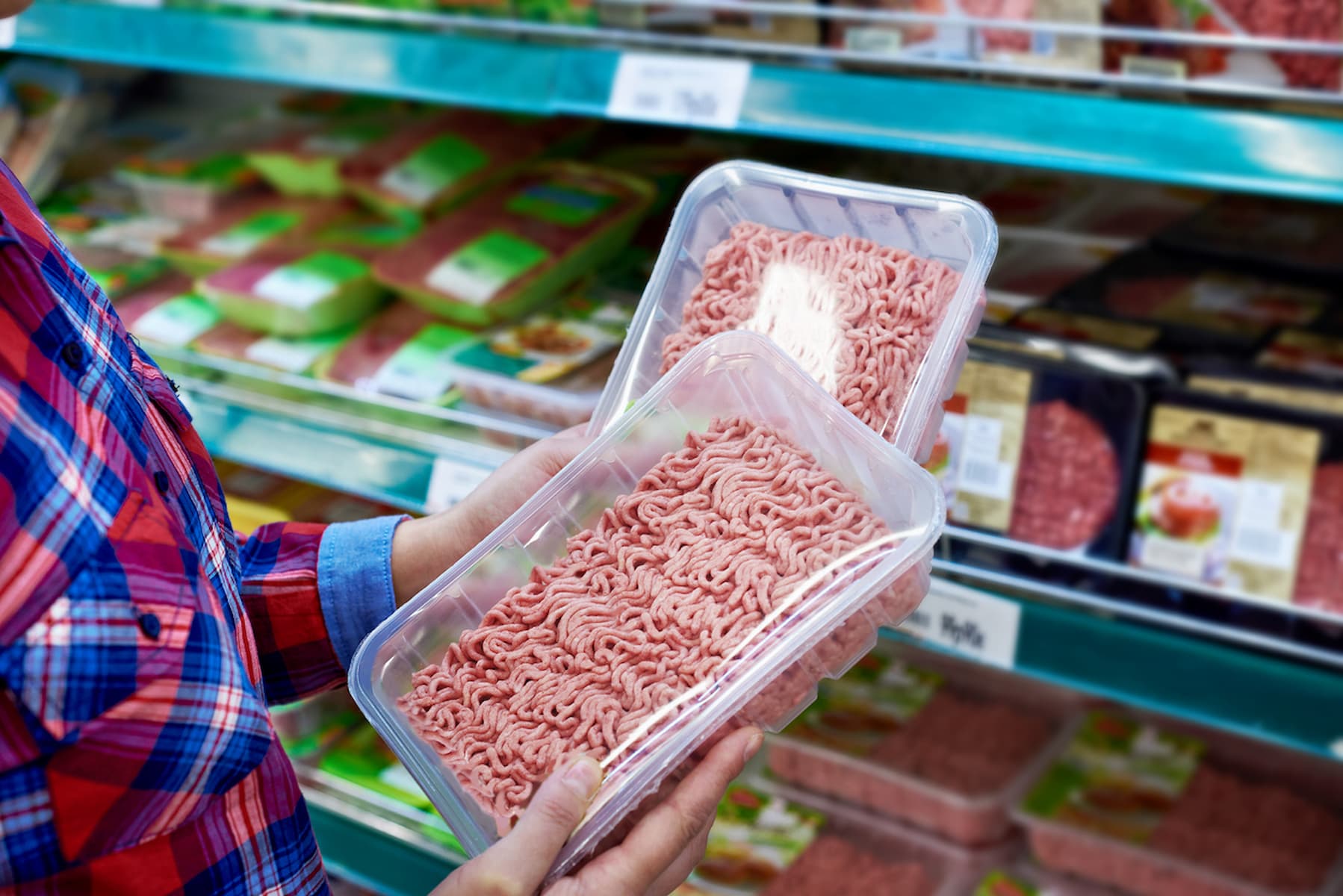 A person in a plaid shirt holds two packs of ground meat while shopping in a grocery store meat aisle. Other packaged meat products are visible on the shelves in the background.