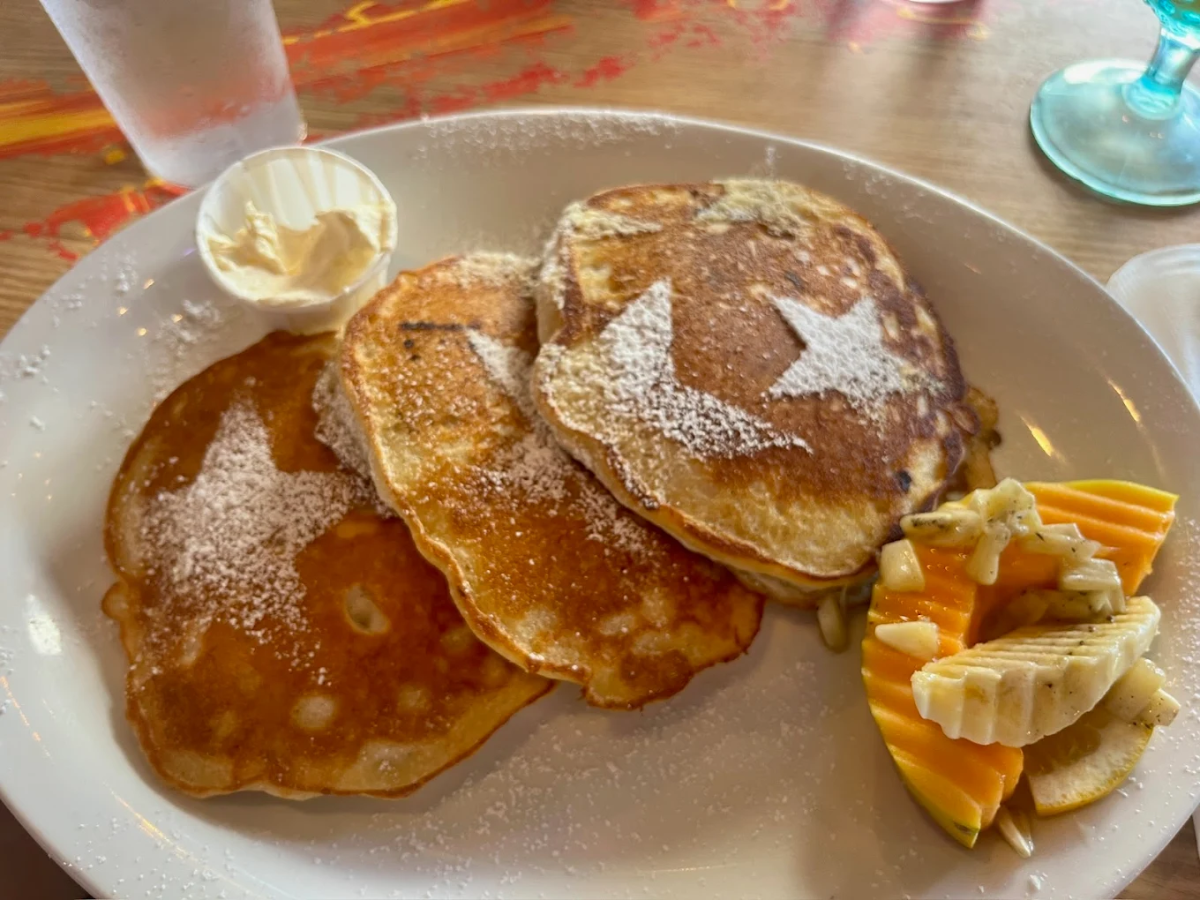 Three pancakes dusted with powdered sugar and decorated with star shapes, served on a white plate with a scoop of butter, sliced bananas, and sliced papaya. A glass of water is in the background.