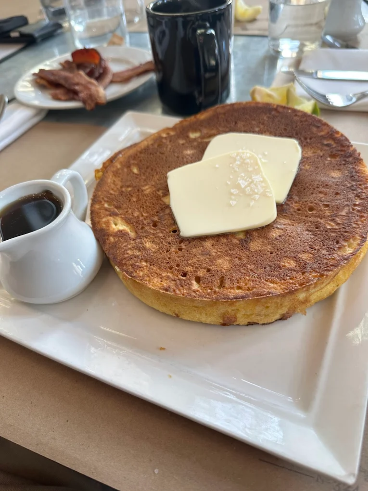 A thick, round pancake topped with two slices of butter and a sprinkle of salt, served on a white plate with a small pitcher of syrup. Bacon and a mug are visible in the background.