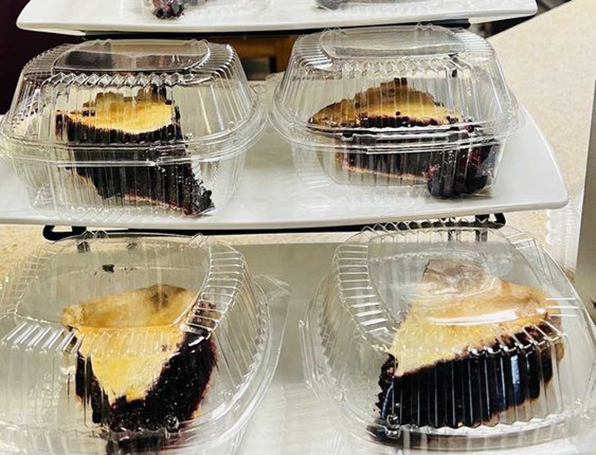 Slices of pie with dark berry filling and golden crust are displayed in clear plastic containers, arranged on white tiered trays.