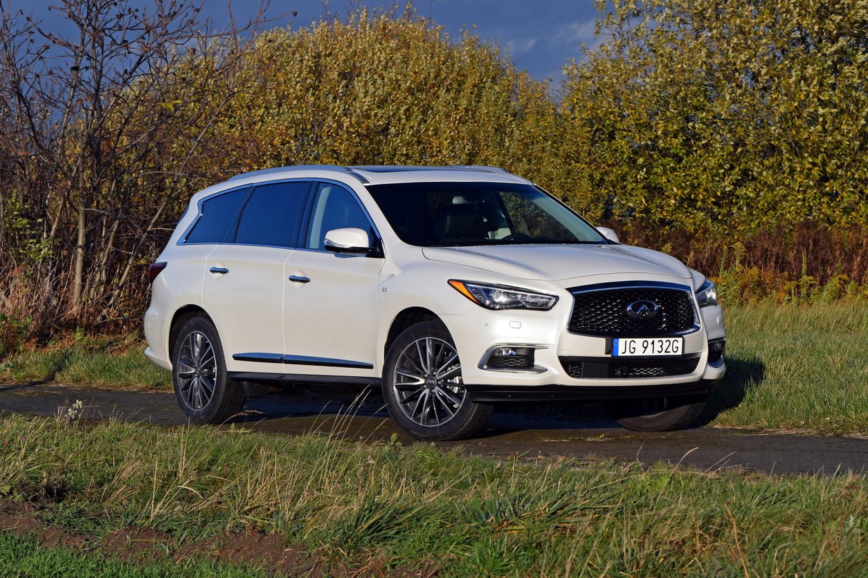 A white Infiniti SUV is parked on a narrow paved road surrounded by grass and trees with autumn leaves under a partly cloudy sky.