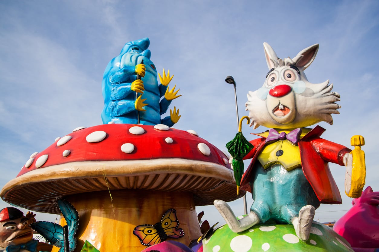 Colorful parade float featuring a large white rabbit in a red jacket and a blue caterpillar, both cartoonish, sitting on oversized mushrooms with polka dots. Bright butterflies and other whimsical decorations surround them.